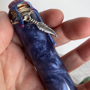 May include: A small, cylindrical glass bottle with a cork stopper, sealed with blue and red wax. The bottle is filled with a swirling, dark blue liquid and adorned with a silver feather charm. The bottle is held in a hand.