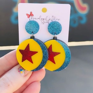 May include: A pair of dangle earrings featuring a blue glitter background with a yellow circle and a red star in the center. The earrings are labeled "Marvelous On Main Street".