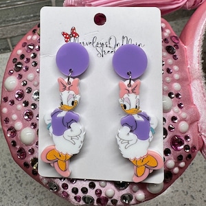 May include: Purple and white dangle earrings featuring a cartoon character with a pink bow. The character is wearing a purple dress and has a yellow beak. The earrings are labeled "Marvelous On Main Street".