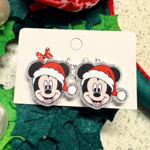 May include: A pair of Mickey Mouse Christmas earrings. Each earring features Mickey's face wearing a Santa hat. The earrings are on a white card with a red bow. The background includes green and red holiday decorations.