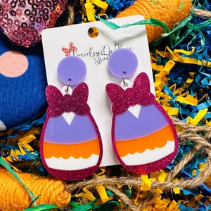 May include: Purple, orange, and white striped egg-shaped earrings with a pink glitter bow. The earrings are made of acrylic and have a shiny finish.