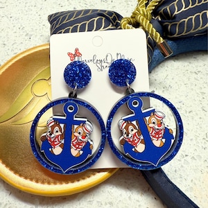 May include: Blue glitter acrylic earrings featuring Chip and Dale in sailor outfits inside a blue anchor design. The earrings have a round blue glitter top and are attached to a white card with text.