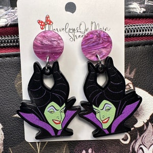May include: Purple and black earrings featuring a cartoon image of Maleficent from Disney's Sleeping Beauty. The earrings have a purple and white swirl design.