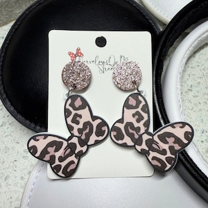 May include: A pair of dangle earrings featuring a butterfly design with a pink and brown leopard print. The earrings have a glittery, round top and are attached to a white card with the text "Marvelous On Main Street".