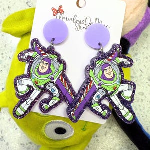 May include: Purple glitter earrings with a cartoon image of Buzz Lightyear from Toy Story. The earrings are shaped like Buzz Lightyear's wings and have a purple glitter background.