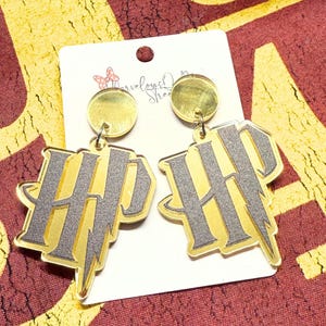 May include: A pair of gold and gray acrylic earrings featuring the letters "HP" with a lightning bolt design. The earrings are hanging from a gold circle stud. The earrings are on a white card with the text "Marvelous Street".