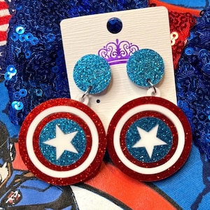 Puede incluir: Un par de pendientes colgantes con un dise&ntilde;o de escudo del Capit&aacute;n Am&eacute;rica rojo, blanco y azul con purpurina. Los pendientes est&aacute;n en una tarjeta blanca con una corona morada.