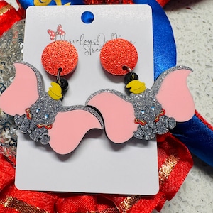 May include: A pair of dangle earrings featuring a red glitter circle with a silver glitter Dumbo the Elephant charm. The elephant has pink ears and a yellow hat.