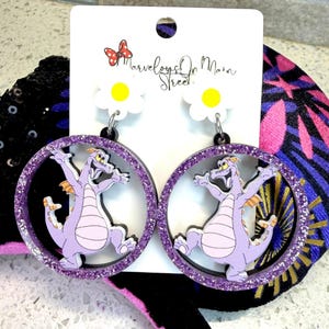May include: A pair of dangle earrings featuring a purple dragon character inside a glittery purple circle. The earrings hang from white daisy-shaped studs with yellow centers. The text "Marvelous on Main Street" is visible on the tag.