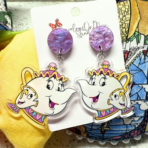 May include: A pair of dangle earrings featuring a cartoon image of Mrs. Potts from Disney's Beauty and the Beast. The earrings have a purple iridescent background and a clear acrylic charm.