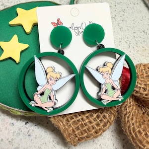 May include: A pair of dangle earrings featuring a green circular frame with a central illustration of a blonde fairy with light blue wings wearing a green dress. The earrings have a green circular stud top and are attached to a white display card.
