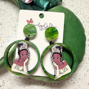 May include: Green hoop earrings featuring a cartoon image of a princess. The earrings have a circular design with a green marbled effect and a black post. The earrings are attached to a white card with the text "Marvelous On Main Street".