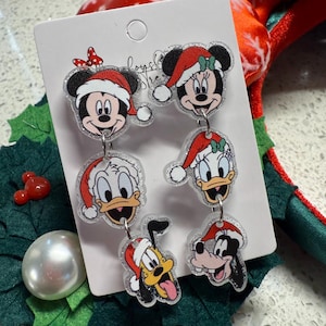 May include: A set of six Christmas-themed dangle earrings featuring Mickey Mouse, Minnie Mouse, Donald Duck, Daisy Duck, Pluto, and Goofy. Each character is wearing a Santa hat. The earrings are attached to a white card.