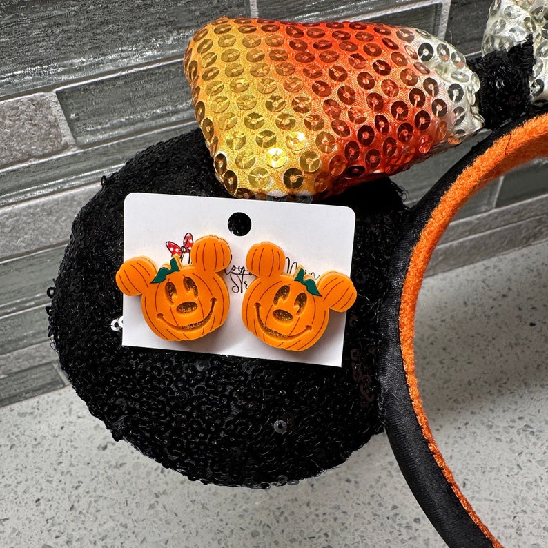 May include: A pair of orange pumpkin-shaped Mickey Mouse ears with a smiling face. The ears are on a black headband with orange trim. The headband is decorated with orange, yellow and white sequins.