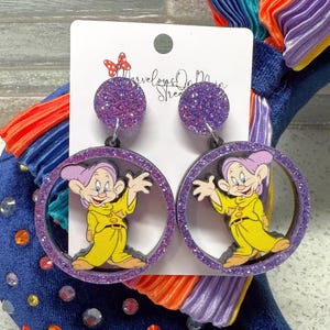 May include: Purple glitter earrings with a round, purple glitter charm and a round, purple glitter frame with a cartoon image of a dwarf from Snow White.