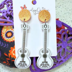 May include: A pair of white guitar-shaped earrings with a skull design and orange glitter accents. The earrings are hanging from a white card with the text "Treloar On Main Street".