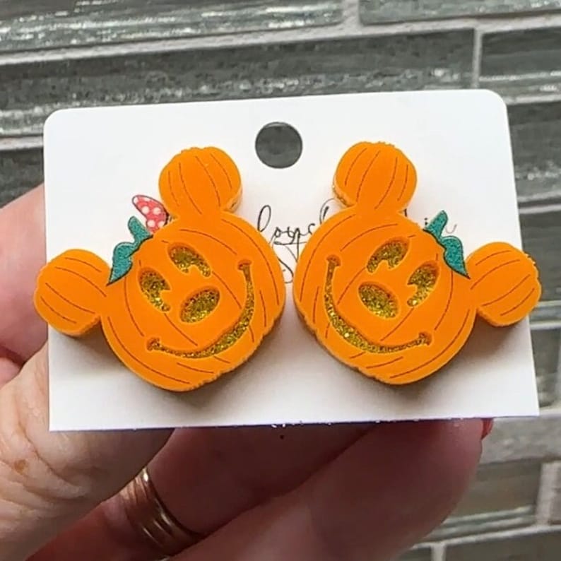 May include: A pair of orange pumpkin-shaped earrings with a Mickey Mouse face design. The earrings have a glitter finish and are perfect for Halloween.