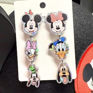 May include: A pair of dangling earrings featuring Mickey Mouse, Minnie Mouse, Daisy Duck, Donald Duck, Goofy, and Pluto. The earrings are made of clear acrylic with a glittery, iridescent finish. Each character is depicted in full color.