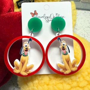 May include: A pair of dangle earrings featuring a cartoon image of Pluto inside a red circle. The earrings have green circular studs and are attached to a white card with the text "Marveloys On Main Street".