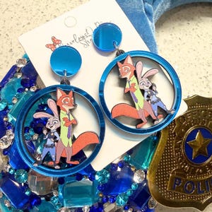 May include: Blue acrylic earrings featuring characters from Zootopia. Each earring has a circular frame with a fox and a bunny. The earrings have a blue disc at the top and are on a white card with a butterfly logo.