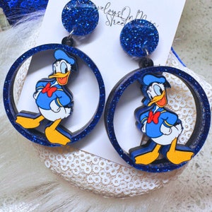 May include: Blue glitter dangle earrings with a round cutout featuring a cartoon image of Donald Duck. The earrings are made of wood and have a black backing.