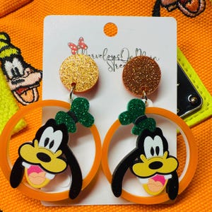May include: Orange hoop earrings with a cartoon image of Goofy's face inside the hoop. The earrings have a gold and copper glitter accent.