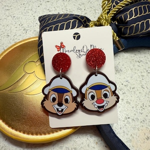 May include: A pair of novelty earrings featuring Chip and Dale characters. The earrings have red glitter circle tops and dangle down to the cartoon faces. The earrings are on a white card with the text "Marvelous on Main Street".