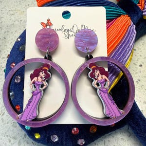 May include: A pair of purple hoop earrings featuring a cartoon image of a woman in a purple dress. The earrings have a glittery finish and are set against a white card with text. The earrings are part of a larger collection of accessories.