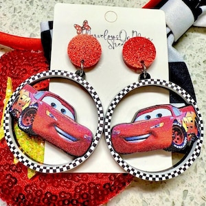 May include: A pair of red glitter-topped earrings featuring a cartoon car design within a black and white checkered hoop. The earrings are attached to a white card with text and a red sequined background.