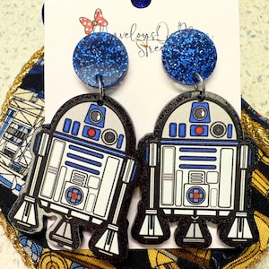 May include: A pair of blue glitter and white R2-D2 themed dangle earrings. The earrings are made of acrylic and have a blue glitter top and a white R2-D2 character charm.