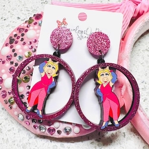 Peut inclure: Boucles d'oreilles rondes en paillettes roses avec un personnage rose de Miss Piggy &agrave; l'int&eacute;rieur d'un cercle plus grand. Les boucles d'oreilles sont sur un fond blanc.