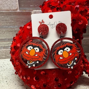 May include: Red glitter earrings with a red and black cartoon image of a furry red monster with a large mouth and a spiked collar. The earrings are on a white card with the text "Weloveys Designs Street".