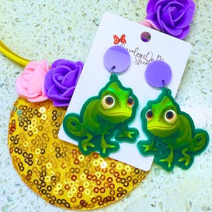 May include: A pair of green chameleon earrings with purple accents. The earrings are on a white card with the text "Revelog On My Street".