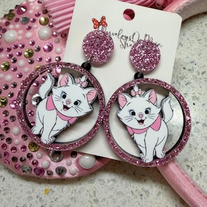 May include: Pink glitter earrings featuring a white cartoon cat with a pink bow and collar. The earrings have a circular design with a pink glitter border and a black outline. The text "Marvelous Main Street" is on the tag.