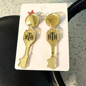 May include: Gold-colored key-shaped earrings with a circular top and a key design. The key design has the letters "HTH" in black. The earrings are hanging on a white card with a red bow and text in the background.