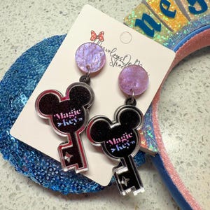 May include: A pair of dangle earrings featuring a Mickey Mouse key design. The earrings have a black glitter outline with the words "Magic Key" in the center. The top of the earrings are round, with a purple and white marbled effect.