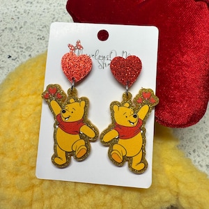 May include: A pair of dangle earrings featuring a yellow cartoon bear with a red scarf, holding hearts. The earrings have red glitter heart-shaped tops. The earrings are on a white card with text.