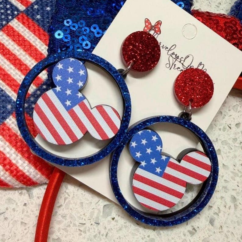 May include: Patriotic earrings featuring a Mickey Mouse design with the American flag. The earrings have a blue glittery border and red glitter accents. The design is set against a white background with a red and blue headband.