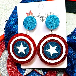 May include: A pair of dangle earrings featuring a red, white, and blue Captain America shield design with glitter. The shield has a white star in the center. The earrings are attached to a blue glitter circle.