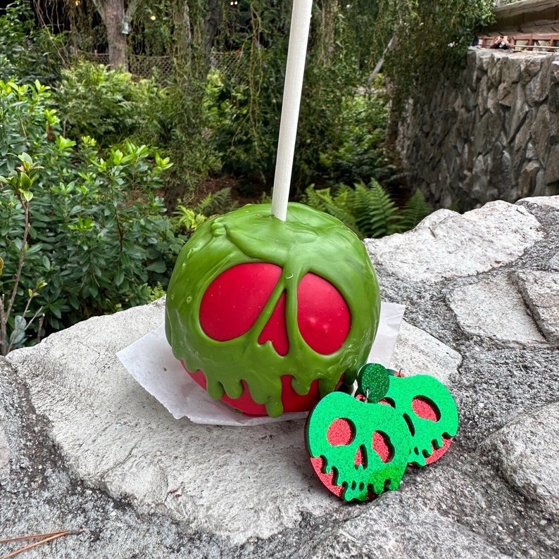 Poison Apple earrings in red and green acrylic, inspired by Snow White. Disney villain earrings displayed on card with Halloween props. Close-up of dripping apple design on lightweight acrylic earrings. Model wearing Poison Apple earrings for spooky Disney outfit. Poison Apple earrings styled with villain-themed accessories and park-ready outfit.