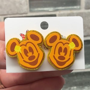 May include: A pair of gold glitter Mickey Mouse earrings with a waffle pattern and a yellow honey drizzle on top.
