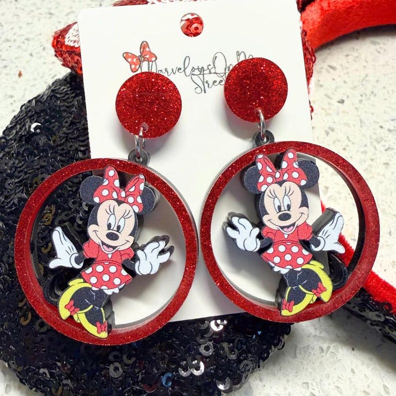 K&ouml;nnte beinhalten: Rote Minnie Mouse Ohrringe mit Glitzer. Jeder Ohrring hat einen roten Glitzerkreis mit einer Minnie Mouse Figur. Die Minnie Mouse Figur tr&auml;gt ein rotes Kleid mit wei&szlig;en Punkten und eine passende Schleife. Die Ohrringe sind an einem roten Glitzerkreis befestigt.
