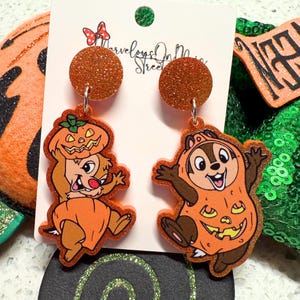 May include: A pair of Halloween-themed dangle earrings. Each earring features a cartoon character in an orange pumpkin costume. The earrings have a glittery orange circle at the top and are attached to a white card with the text "Marvelous On Main Street".