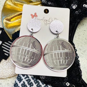 May include: A pair of silver-toned Death Star earrings. Each earring features a detailed gray and white graphic of the Death Star, suspended from a small, glittery silver circle. The earrings are displayed on a white card with the text "Marvelous Dreams Street".