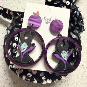 May include: A pair of dangle earrings featuring a circular purple frame and a round purple stud. Inside each circular frame is a graphic illustration of a character with black horns, a high purple collar, and light green skin. The earrings are attached to a white display card.
