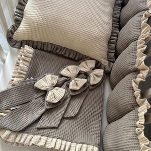 Könnte beinhalten: Ein Babybett-Set aus grauem Waffelstoff. Das Set beinhaltet ein Kissen, Stoßfänger und eine Decke mit dekorativen Schleifen. Die Kanten sind mit einer kontrastierenden beigen Rüsche versehen.