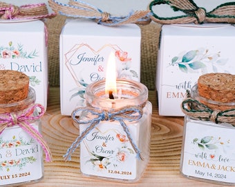 May include: Four small glass jars with cork lids and labels. Each jar contains a small white candle and has a different wedding-themed label with the couple's names and wedding date. The jars are arranged on a wooden slice with hessian fabric in the background.