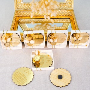 May include: Six small white boxes with gold trim, each containing a round, gold-colored, mirrored, wooden coaster with a decorative edge. The coasters have Arabic calligraphy on them and are decorated with dried flowers. Three additional coasters are shown outside of the boxes, one with a magnet on the back.
