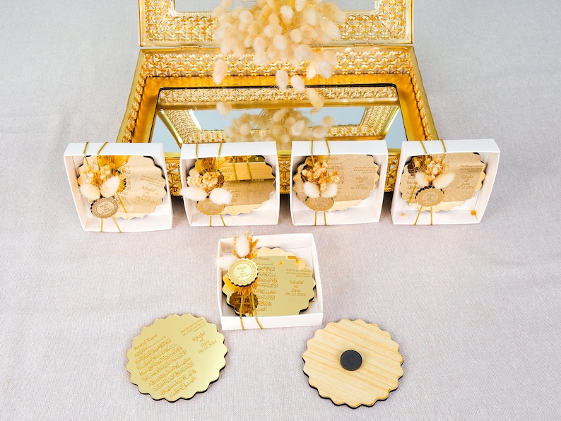 May include: Six small white boxes with gold trim, each containing a round, gold-colored, mirrored, wooden coaster with a decorative edge. The coasters have Arabic calligraphy on them and are decorated with dried flowers. Three additional coasters are shown outside of the boxes, one with a magnet on the back.