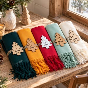 May include: Five folded rectangular scarves in dark green, mustard yellow, red, light green, and cream, each featuring a fringed edge and a wooden Christmas tree-shaped ornament tied with a ribbon, personalized with names like Emma and Lucy.